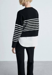 Gestreifter Pullover in Schwarz und Weiß mit Rundhalsausschnitt, der ein kurzes Design über einem längeren weißen Hemd bietet, kombiniert mit schwarzen Hosen.