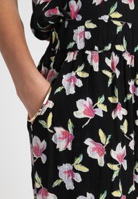 Main dans la poche d'une robe noire avec motif floral rose, blanc et jaune et taille froncée, portant un bracelet en or.