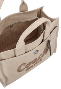 Beige canvas tote bag with a logo, dual handles, zippered pocket, and interior with fabric lining. Features metallic hardware accents.