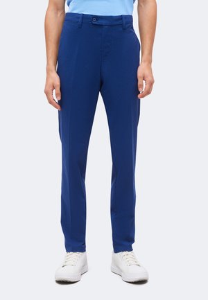 Homme portant un pantalon bleu ajusté avec fermeture à boutons et des baskets blanches, debout devant un fond clair uni.