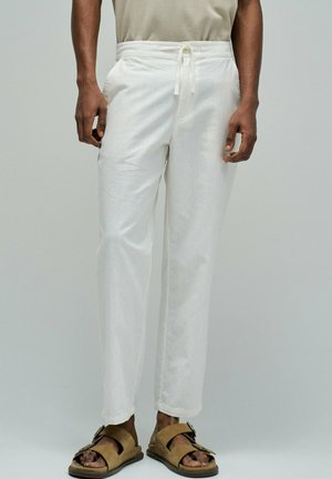 Hombre con pantalones blancos con cordón y sandalias marrones con hebilla, sobre un fondo claro liso.