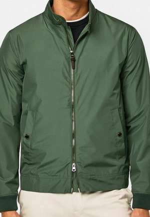 Mężczyzna w zielonej kurtce na zamek z zapinanymi na guziki bocznymi kieszeniami i ściągaczami przy mankietach, noszący pod nią czarno-białą koszulę.