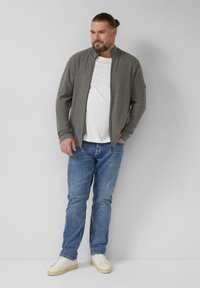Grauer Zip-up-Cardigan mit hohem Kragen, getragen über einem weißen T-Shirt, kombiniert mit hellblauen Jeans und weißen Turnschuhen. Einfacher, lässiger Look.