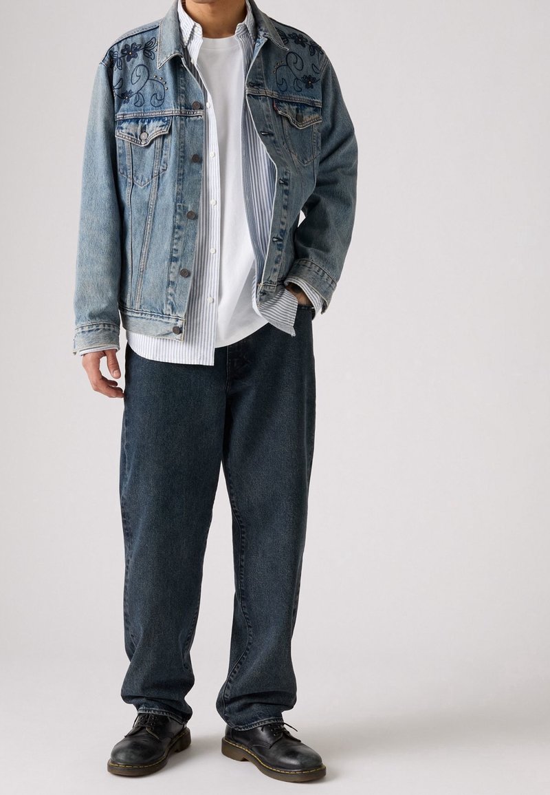 Personne portant une veste en jean brodée par-dessus une chemise rayée et un t-shirt blanc, un jean ample foncé et des chaussures en cuir noires, debout devant un fond uni.