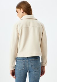 Beige kurz geschnittene Jacke mit Reverskragen, langen Ärmeln und Druckknopfmanschetten, kombiniert mit blauen Jeans. Glatte Textur und taillierte Passform.