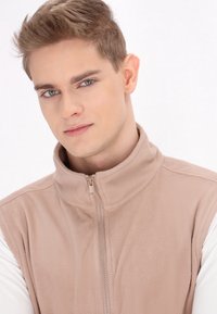 Gilet in pile color beige chiaro con colletto alto e zip verticale. Indossato sopra una camicia bianca a maniche lunghe. Tessuto morbido, design casual.