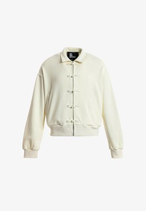 Chaqueta off-white con cremallera, cuello ancho, puños y dobladillo acanalados, con cinco cierres decorativos en forma de nudos de tela en la parte delantera.