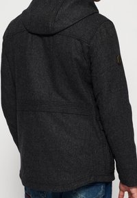 Donkergrijze jas van een wolmix met een capuchon, met een getextureerde afwerking en een zijflap op de linkerarm. Eenvoudig ontwerp zonder zichtbare sluitingen.