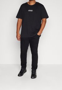 Camiseta negra con la inscripción "COSMIC MYSTERY" en blanco, combinada con pantalones negros y zapatillas negras de caña alta, sobre un fondo neutro.