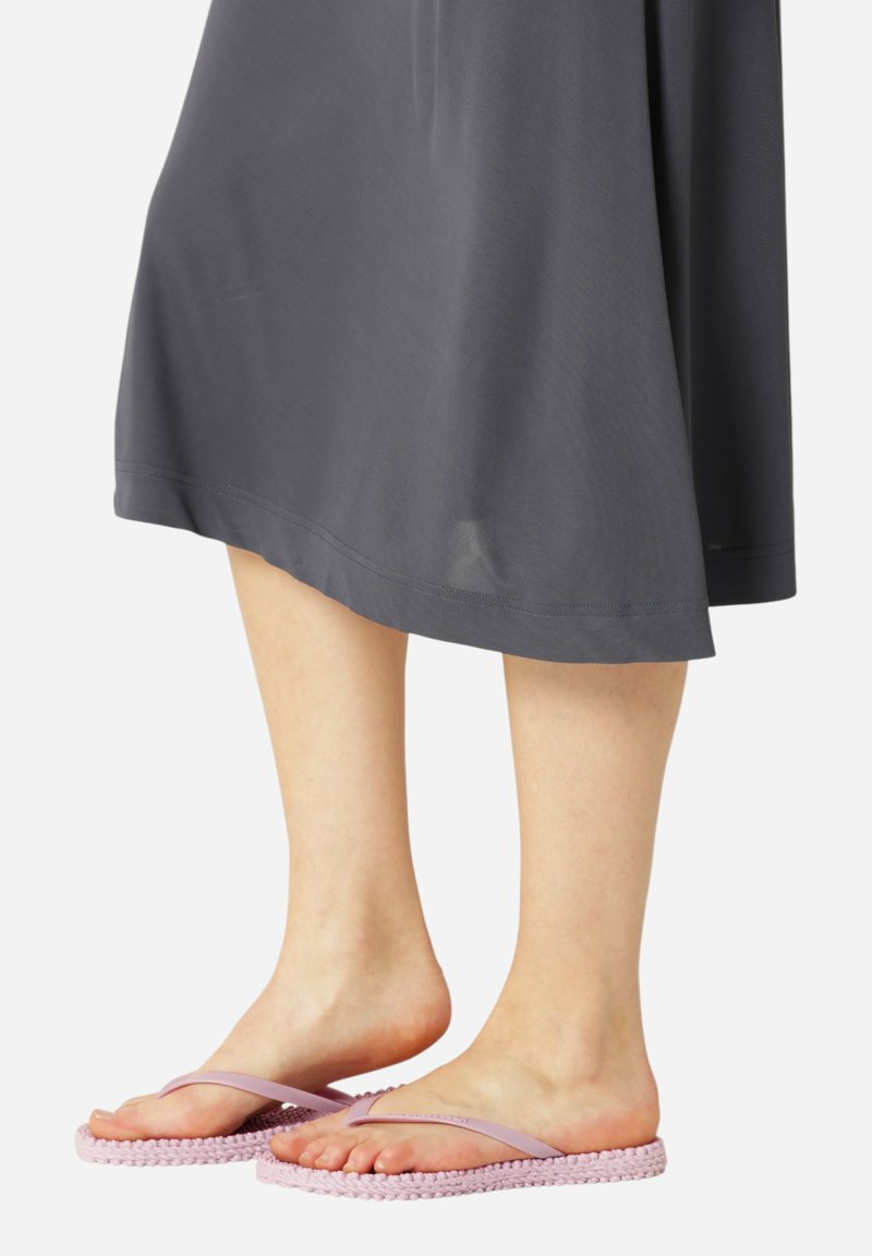 Blote voeten in lichte paarse flip-flops met gehaakte zolen, bedekt door een grijze rok tot de knie tegen een witte achtergrond.