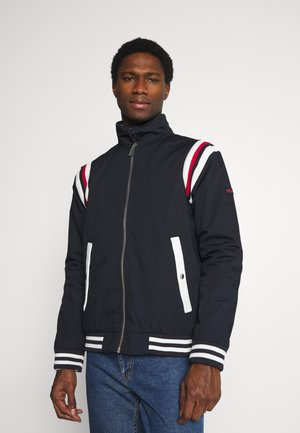 Veste zippée marine avec un col montant, fermeture éclair blanche et bandes rouges et blanches sur les épaules. Elle possède deux poches latérales blanches et des poignets rayés.