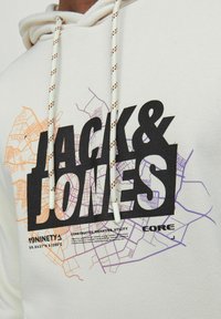 Cremefarvet hoodie med et stort sort "JACK & JONES" logo, prydet med lilla og orange kortmønstre. Inkluderer mønstrede snore og en blød tekstur.