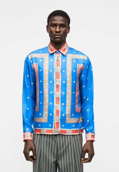 Bărbat care poartă o cămașă din satin albastră cu modele decorative roșii, bej și mici motive albe, asortată cu pantaloni în dungi verticale alb-negru.