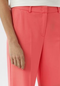 Jasne koralowe spodnie wykonane z gładkiego materiału o dopasowanym kroju, z przodu plisami i kieszeniami bocznymi, na zdjęciu z częściowo widoczną ręką.