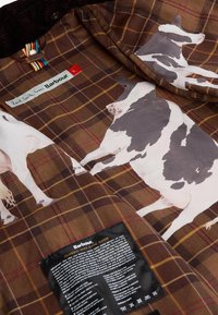 Tecido de padrão xadrez castanho com ilustrações de vacas, uma etiqueta de marca e instruções de cuidados em preto sobre um fundo contrastante.