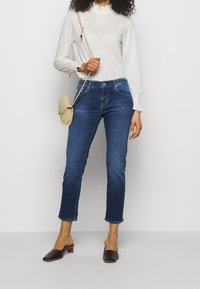 Mörkblå denimjeans med slim fit och kort längd, kombinerade med en vit långärmad blus med en VOLANGKRAGE och manchetter.