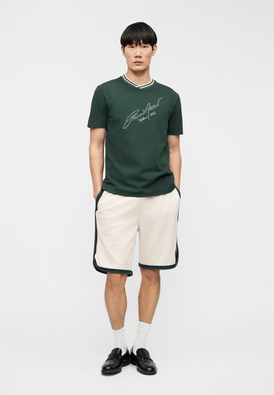 Σκούρο πράσινο βαμβακερό μπλουζάκι με λευκό περιθώριο και λογότυπο, συνδυασμένο με κρεμ σορτς που διαθέτουν λεπτομέρειες σε σκούρο πράσινο και πλαϊνές τσέπες. Μαύρα μοκασίνια.