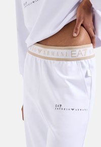 Bele trenirke z zlato-bež elastičnim pasom, ki nosi blagovno znamko "EA7 EMPORIO ARMANI", in stranskim žepom. Gladka tekstura.