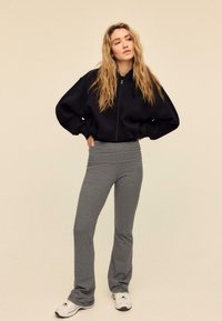 Schwarzer Kapuzenpullover mit Reißverschluss und voluminösen Ärmeln, kombiniert mit grauen, gestreiften Schlaghosen und weißen Sneakers. Weiche Stoffe mit strukturierten Mustern.