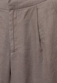 Primer plano de unos pantalones de lino marrón claro que muestran la textura de la tela, la cintura, los pliegues y los detalles de la costura lateral.