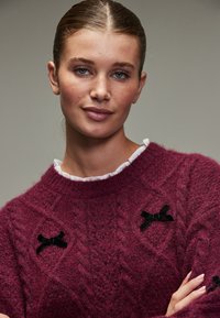 Bordeauxroter Strickpullover mit Zopfmuster, verziert mit schwarzen Samtschleifen und einem weißen Rüschenkragen. Weiches, strukturiertes Material.