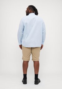 Chemise bleu clair à manches longues avec des poignets à boutons, associ�ée à un short roulé beige. Des chaussures noires et des chaussettes complètent la tenue.