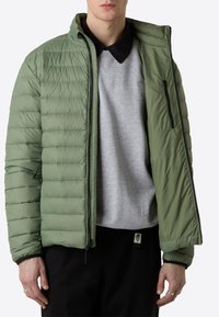 Giacca a vento leggera di colore verde oliva, con cuciture orizzontali, tasche con zip e colletto alto, indossata sopra un felpa grigia.