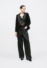 Zwarte blazer met textuur en satijnen revers, gecombineerd met een glanzende zwarte top en wijde leren broek. Model staat op een neutrale achtergrond.