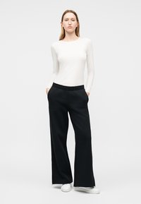 Nő áll fekete bő szárú nadrág zsebében tartott kézzel, fehér hosszú ujjú felsőt és fehér tornacipőt visel, semleges háttér előtt.