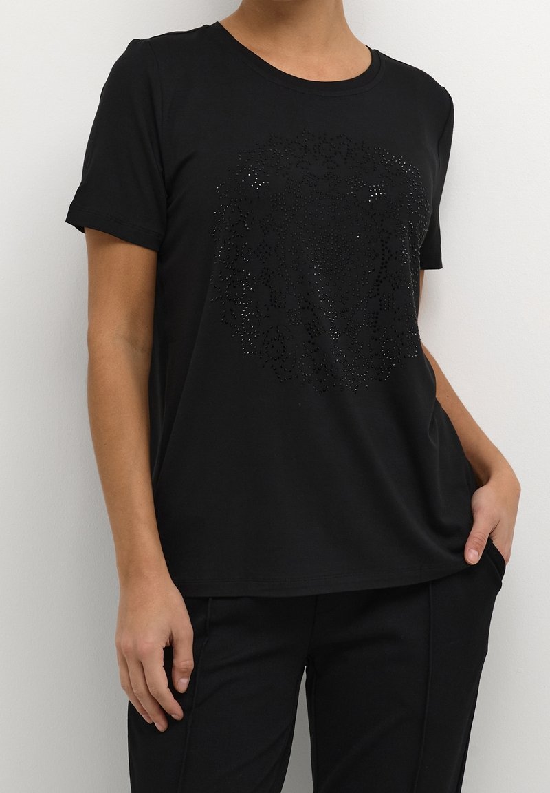 Femme portant un t-shirt noir à manches courtes avec un motif floral subtil, brillant et texturé sur la poitrine, associé à un pantalon noir.