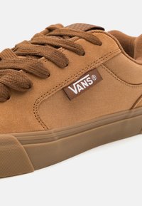 Brązowe płócienne trampki z pasującymi sznurówkami, wzmocnionym palcem, teksturowaną podeszwą i białą łatą z logo z boku. Casualowy design z wyściełanym kołnierzem.