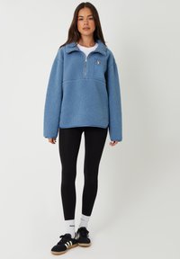 Lys blå fleece-pullover med halvlukning og brystlomme. Har brede ærmer og struktureret stof, parret med sorte leggings og sneakers.