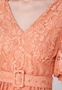 Robe en dentelle pêche avec un décolleté en V, des manches courtes et une taille ceinturée doté d'une boucle rectangulaire et d'accents dorés. Motif floral sur l'ensemble.