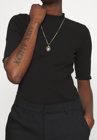 Svart ribbad topp med korta, volangärmar, bärs med högmidjade mörka byxor. Guldpendelhalsband vilar på U-ringningen.