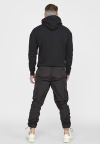Sweat à capuche noir à coupe décontractée, associé à un pantalon cargo de jogging noir avec plusieurs poches et des poignets élastiques. Baskets blanches visibles.