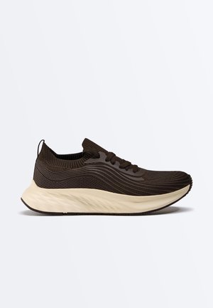 Sötétbarna kötött futócipő hullámos mintával, fűzős kialakítással, párnázott szegéllyel és vastag, fehér párnázott talppal.