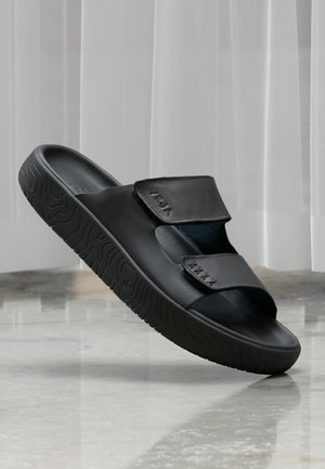 Black slide sandals with a textured rubber sole and dual adjustable velcro straps. Smooth upper material with embossed branding detail.
