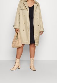 Kvinna som bär en beige trenchcoat över en svart klänning, håller en beige mjuk handväska och har på sig beiga snörstövletter med klack.