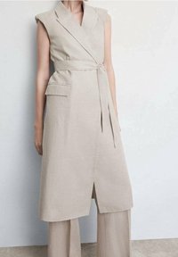 Beżowa lniana sukienka bez rękawów, z kopertowym krojem, paskiem w talii, boczną kieszenią i ostrym kołnierzem. Na dole znajduje się przedni rozporek, który zapewnia większą swobodę ruchów.