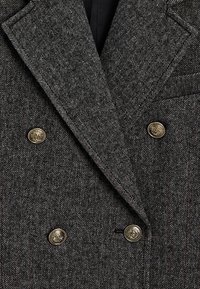 Grijze houndstooth blazer met een dubbelgebreid ontwerp, opstaande revers en metalen knopen. De stof heeft een getextureerde, geweven uitstraling.