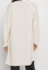 Lång, öppen stickad cardigan i krämfärg. Har en avslappnad passform, ribbad textur och sidofickor för ökad rörelsefrihet.