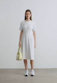 Λευκό βαμβακερό φόρεμα t-shirt με κοντά μανίκια και πράσινες λεπτομέρειες, με εφαρμοστό μπούστο και φαρδιά φούστα. Συνδυασμένο με κίτρινη τσάντα και αθλητικά παπούτσια.