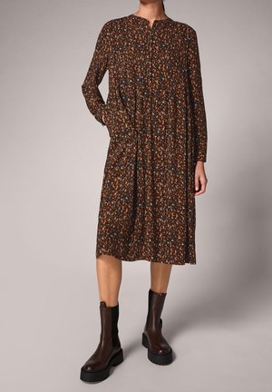 Femme portant une robe fleurie foncée longueur genoux avec manches longues et des bottines marron à plateforme, debout devant un fond uni.