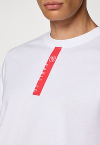 Hvid bomulds T-shirt med rund hals, der har et lodret rødt grafisk strib og tekst, der inkluderer "90" og "PARIS SG."