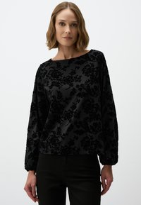 Zwarte flonkerende fluwelen blouse met een bootnek, lange pofmouwen en een gestructureerd patroon. Gecombineerd met zwarte broek.