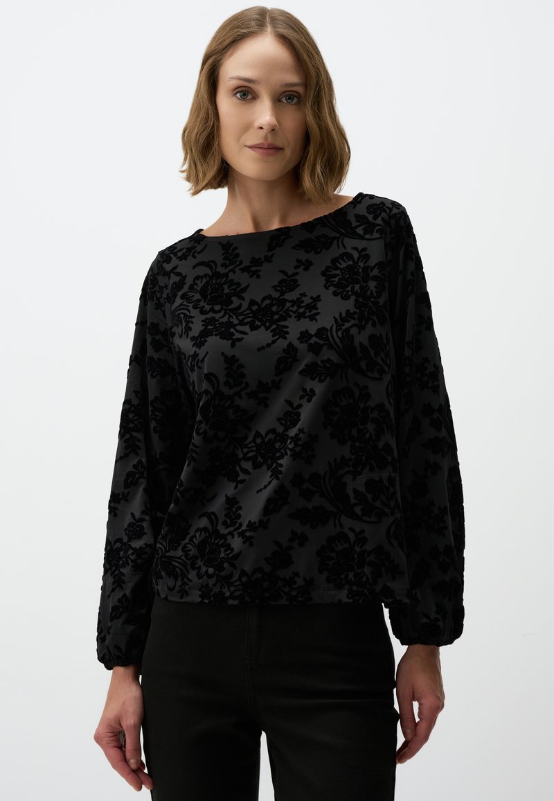 Zwarte flonkerende fluwelen blouse met een bootnek, lange pofmouwen en een gestructureerd patroon. Gecombineerd met zwarte broek.
