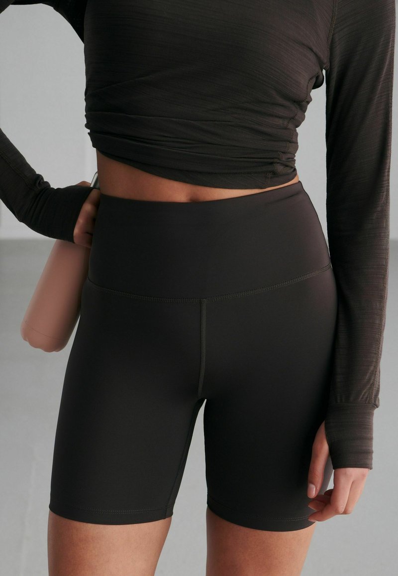 Zwarte high-waisted sportshorts gemaakt van rekbare stof, met flatlocknaden en een gladde textuur. Hand die een waterfles vasthoudt.