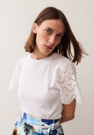 Femme aux cheveux châtains mi-longs portant un haut blanc avec des manches en dentelle ajourée et une jupe fleurie colorée sur un fond uni.