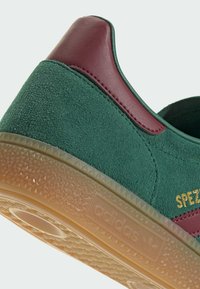 Zapatillas de ante verde con acentos de cuero granate, suela de goma texturizada y detalle de marca en dorado, presentando una forma redondeada y un diseño minimalista.