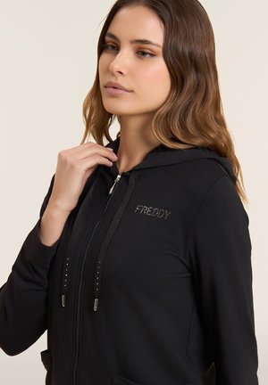Joven con el cabello largo y castaño que lleva una sudadera negra con cremallera y el logo "FREDDY" en pedrería, ajustando el cordón de la capucha sobre un fondo claro.
